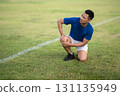 An asian man in blue t shirt is kneeling and holding his knee on grass field. Pain or injury. 131135949