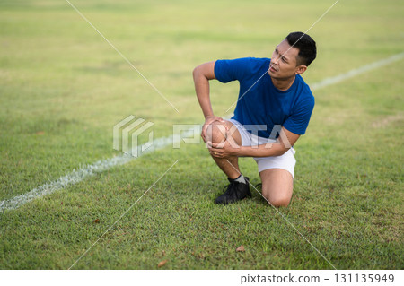 An asian man in blue t shirt is kneeling and holding his knee on grass field. Pain or injury. 131135949