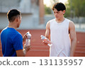 Two asian athlete men are looking at their water bottles while talking together on the racetrack in sports stadium. 131135957