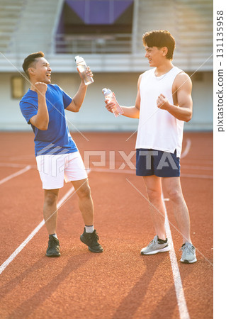 Two asian men are holding water bottles while talking together on the racetrack in sports stadium. 131135958