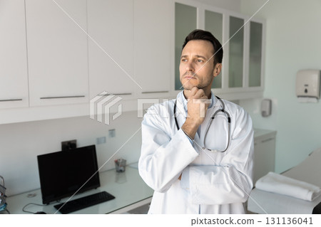 Serious male doctor standing in examining room deep in thought Serious male doctor standing in examining room deep in thought 131136041