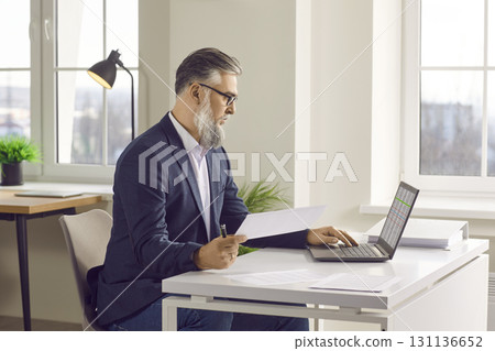 Senior male financial accountant sitting at office desk and working on laptop computer 131136652