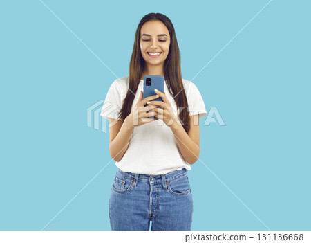Happy brunette girl in white t-shirt is reding looking at smartphone screen on blue background. 131136668