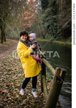 smiling woman in yellow coat holding her small child in hands standing in the park next to the river smiling woman in yellow coat holding her small child in hands standing in the park next to the river 131137464