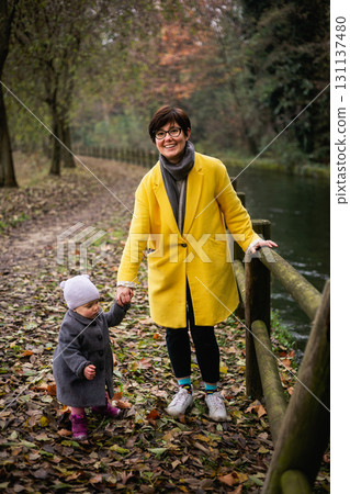 woman in yellow coat and small child in grey coat walking together in the park next to the river 131137480
