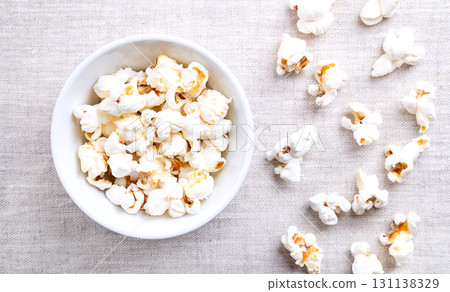 Popcorn, popped corn in a white bowl on linen. Seeds of the Zea mays variety everta, expanded and puffed up, after heated. Snack food, also called popcorns and pop-corn. Close-up, from above. Photo Popcorn, popped corn in a white bowl on linen. Seeds of the Zea mays variety everta, expanded and puffed up, after heated. Snack food, also called popcorns and pop-corn. Close-up, from above. Photo 131138329