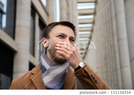 Caucasian young man stands in urban area filled with tall buildings. He is laughing and covering his mouth while wearing stylish coat and scarf. manager, office worker, businessman, lawyer portrait Caucasian young man stands in urban area filled with tall buildings. He is laughing and covering his mouth while wearing stylish coat and scarf. manager, office worker, businessman, lawyer portrait 131138334