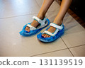 Feet adorned with white sandals are encased in bright blue plastic covers, showcasing a playful precaution against wet flooring during rainy weather, adding a touch of fun to indoor activities. 131139519