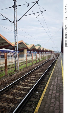 Empty railway tracks at a train station with patterned roofs and electric lines under a cloudy sky 131139539