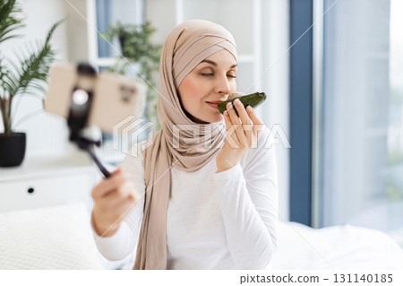 Young Muslim woman in beige hijab holding smartphone on tripod sharing skincare tips using avocado. She discusses natural beauty methods while creating engaging and insightful content in a cozy home Young Muslim woman in beige hijab holding smartphone on tripod sharing skincare tips using avocado. She discusses natural beauty methods while creating engaging and insightful content in a cozy home 131140185