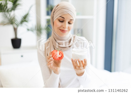Young Muslim woman wearing beige hijab holding fresh tomato and skincare cream while smiling. Daylight ambience with indoors natural decor conveys skincare routine concept. Young Muslim woman wearing beige hijab holding fresh tomato and skincare cream while smiling. Daylight ambience with indoors natural decor conveys skincare routine concept. 131140262