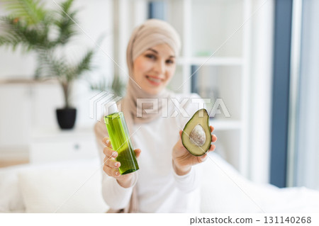Focused view on Muslim adult woman with turban in modern room holding half avocado and green skincare oil bottle, representing organic skincare and health-conscious lifestyle 131140268
