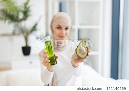 Focused view on Muslim adult woman with turban in modern room holding half avocado and green skincare oil bottle, representing organic skincare and health-conscious lifestyle Focused view on Muslim adult woman with turban in modern room holding half avocado and green skincare oil bottle, representing organic skincare and health-conscious lifestyle 131140269