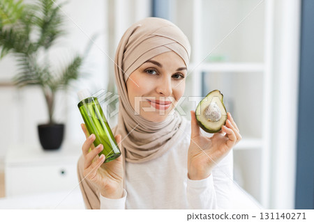 Young Muslim woman in beige headscarf holding green bottle and avocado for natural beauty skincare promotion. Image promotes concepts of organic skincare natural beauty routines and holistic wellness. 131140271