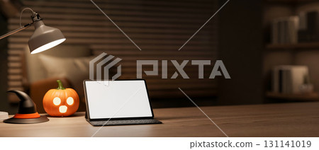 White screen tablet with halloween pumpkin lantern and witch hat under lamp on table in a dark room. 131141019