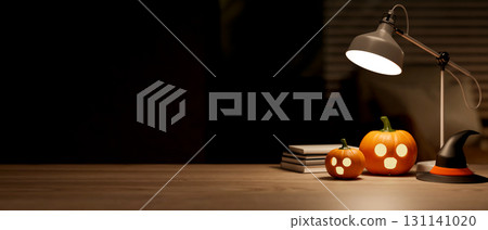 Glowing halloween pumpkin lantern aside witch hat and books under lamp on wooden table in dark room. 131141020