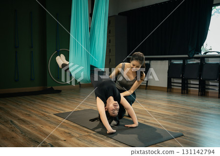 Fitness instructor assisting young woman practicing aerial yoga inversion pose using hammock in studio 131142794