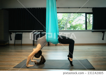 Young woman exercising with aerial yoga hammock, stretching body in advanced backbend posture Young woman exercising with aerial yoga hammock, stretching body in advanced backbend posture 131142805