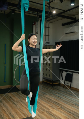 Teenage girl practicing aerial yoga indoors, balancing with fabric hammock support in fitness studio 131142818