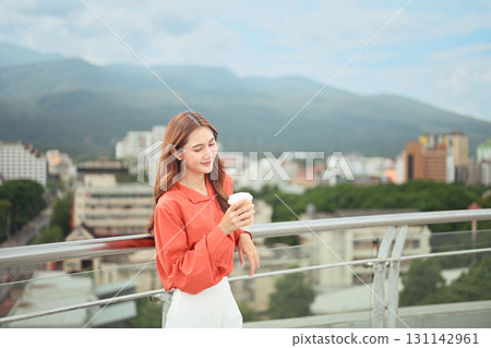Relaxed businesswoman holding takeaway cup while standing on rooftop with city and mountain view 131142961