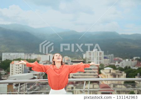 Cheerful woman standing on rooftop with arms wide open, smiling joyfully against cityscape and mountain view Cheerful woman standing on rooftop with arms wide open, smiling joyfully against cityscape and mountain view 131142963