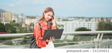 Stressed young businesswoman working on laptop outdoors with cityscape in the background Stressed young businesswoman working on laptop outdoors with cityscape in the background 131143011