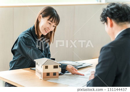 A woman in work clothes explaining and proposing her own home while looking at a house model and blueprints A woman in work clothes explaining and proposing her own home while looking at a house model and blueprints 131144437