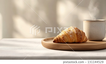 A delicious croissant rests on a wooden plate beside a steaming cup, creating a cozy atmosphere in the sunlit kitchen A delicious croissant rests on a wooden plate beside a steaming cup, creating a cozy atmosphere in the sunlit kitchen 131144529