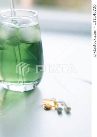 Close-up of chlorophyll iced drink with a glass straw, assorted vitamins in the foreground. Vertical. Close-up of chlorophyll iced drink with a glass straw, assorted vitamins in the foreground. Vertical. 131146720