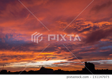 A vibrant sunset with shades of orange, pink, and purple clouds. Silhouettes of buildings are visible in the foreground against the colorful sky. 131147073