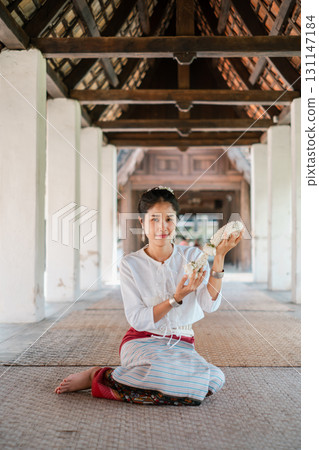 A woman in traditional attire holds a shell, seated in a historic building with wooden beams and tiled roof. 131147184
