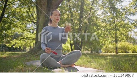A young beautiful white girl doing yoga in the park and meditating in the lotus position among the trees in a clearing on a summer day and using a singing brass bowl A young beautiful white girl doing yoga in the park and meditating in the lotus position among the trees in a clearing on a summer day and using a singing brass bowl 131149067