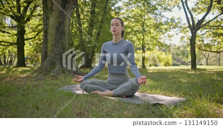 A young beautiful white girl practices yoga in the park and meditates in the lotus position among the trees in a clearing on a summer day 131149150