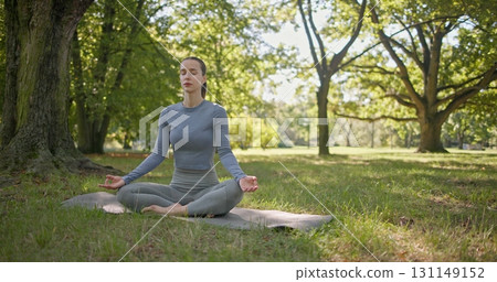 A young beautiful white girl practices yoga in the park and meditates in the lotus position among the trees in a clearing on a summer day A young beautiful white girl practices yoga in the park and meditates in the lotus position among the trees in a clearing on a summer day 131149152