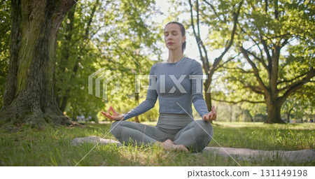 A young beautiful white girl practices yoga in the park and meditates in the lotus position among the trees in a clearing on a summer day A young beautiful white girl practices yoga in the park and meditates in the lotus position among the trees in a clearing on a summer day 131149198