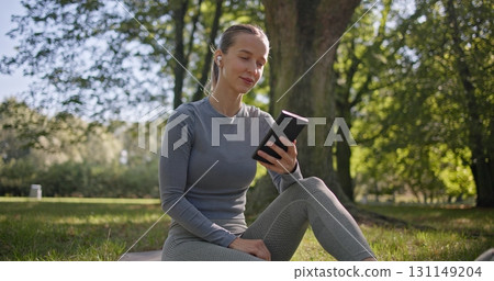 A young beautiful white girl practices yoga in the park and meditates in the lotus position among the trees in a clearing on a summer day and listens to music from her phone with headphones 131149204