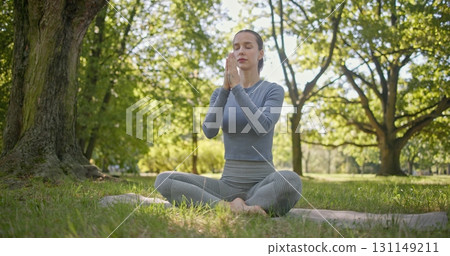 A young beautiful white girl practices yoga in the park and meditates in the lotus position among the trees in a clearing on a summer day 131149211