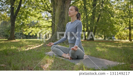 A young beautiful white girl practices yoga in the park and meditates in the lotus position among the trees in a clearing on a summer day A young beautiful white girl practices yoga in the park and meditates in the lotus position among the trees in a clearing on a summer day 131149217