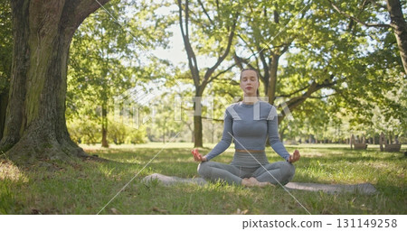 A young beautiful white girl practices yoga in the park and meditates in the lotus position among the trees in a clearing on a summer day 131149258