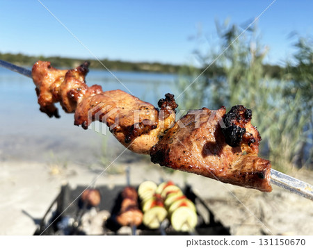 Pork skewer on barbecue grill with smoke and fire against natural lake background. Outdoor cooking in nature, summer picnic and recreation Pork skewer on barbecue grill with smoke and fire against natural lake background. Outdoor cooking in nature, summer picnic and recreation 131150670