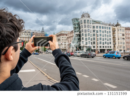 Prague, Czech Republic, August 7, 2023. A young Caucasian man photographs with his cell phone the fascinating three quarter view of the Dancing House: futuristic shapes and modern materials. Prague, Czech Republic, August 7, 2023. A young Caucasian man photographs with his cell phone the fascinating three quarter view of the Dancing House: futuristic shapes and modern materials. 131151599