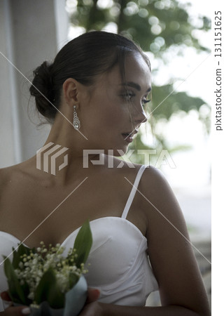 A beautiful girl in a white dress with a bouquet of lilies of the valley stands near the window A beautiful girl in a white dress with a bouquet of lilies of the valley stands near the window 131151625