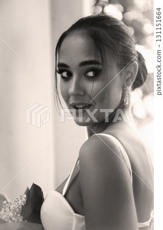 A beautiful girl in a white dress with a bouquet of lilies of the valley stands near the window A beautiful girl in a white dress with a bouquet of lilies of the valley stands near the window 131151664