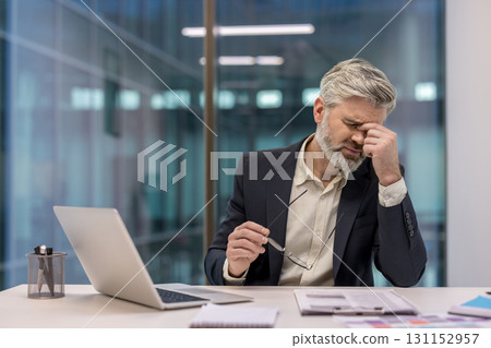Mature businessman experiencing stress and fatigue, rubbing his eyes from a headache while sitting at his desk with a laptop and documents in a modern office environment 131152957