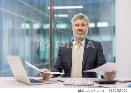 Businessman looking shocked and overwhelmed while reviewing financial papers and dealing with stress, debt, and economic problems in a modern office setup Businessman looking shocked and overwhelmed while reviewing financial papers and dealing with stress, debt, and economic problems in a modern office setup 131152961