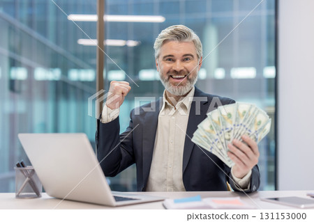 Mature businessman in suit clutching a fan of hundred-dollar bills and pumping his fist at a laptop-covered office desk. Celebrating financial success. Wealth and confident entrepreneurial achievement Mature businessman in suit clutching a fan of hundred-dollar bills and pumping his fist at a laptop-covered office desk. Celebrating financial success. Wealth and confident entrepreneurial achievement 131153000