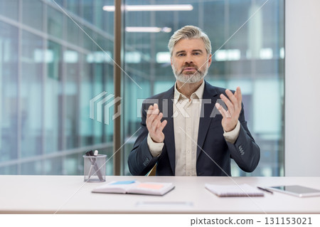 Senior businessman gesturing during an online video conference, discussing business strategies and communicating with clients or colleagues while working from a modern corporate office Senior businessman gesturing during an online video conference, discussing business strategies and communicating with clients or colleagues while working from a modern corporate office 131153021