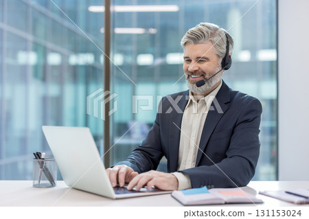 Mature businessman wearing a headset and smiling, actively typing on a laptop, providing customer service or technical support from a modern office environment 131153024
