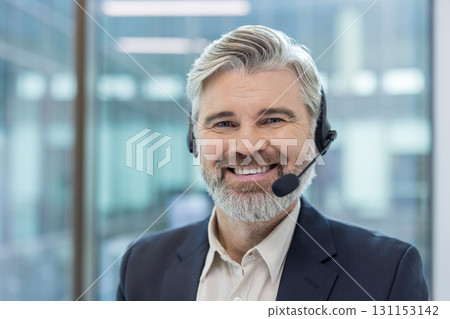 Mature man with a grey beard and headset smiling, representing technical support, customer service, and professional communication in a modern office environment 131153142