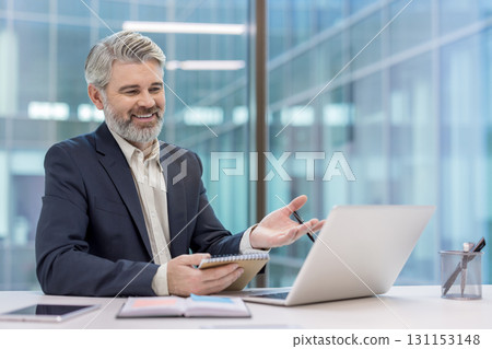 Mature businessman smiling, gesturing, and taking notes during a video conference on his laptop, working in a modern office environment with large glass windows Mature businessman smiling, gesturing, and taking notes during a video conference on his laptop, working in a modern office environment with large glass windows 131153148